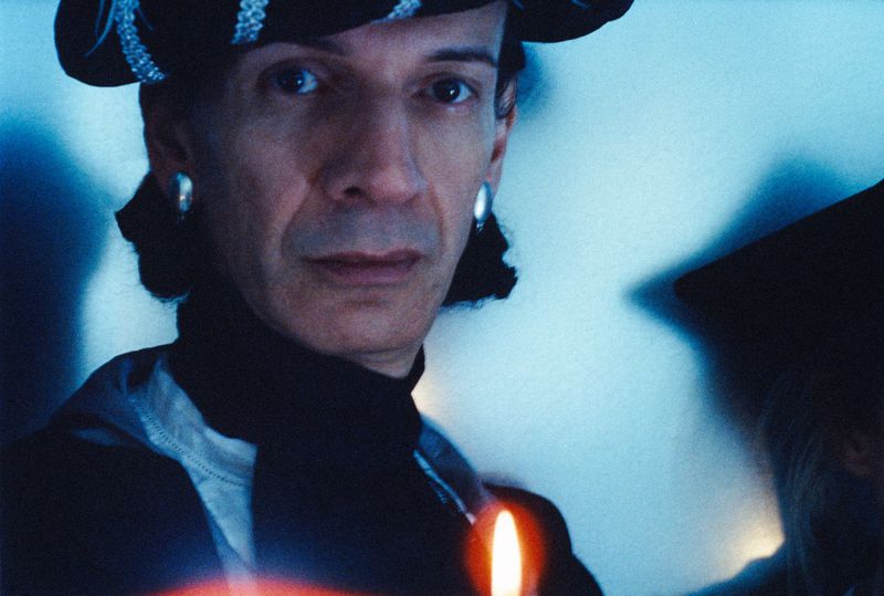 Close-up shot of an adult male with a hat and large earrings, partially obscured face in the background, and a lit candle in the foreground.