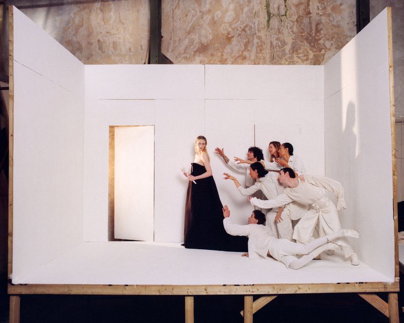A fashion shot inside a makeshift studio shows a woman in a black dress being reached for by a group of people in white outfits.