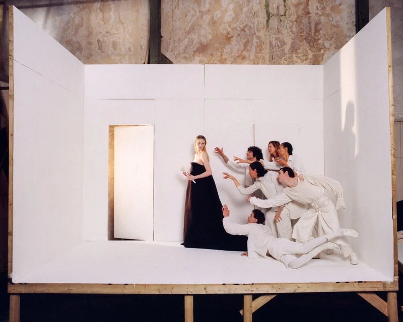 A fashion shot inside a makeshift studio shows a woman in a black dress being reached for by a group of people in white outfits.