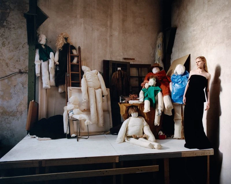 Fashion shoot featuring a blonde woman in a black dress standing next to a tableau of padded mannequin-like figures in an industrial space.