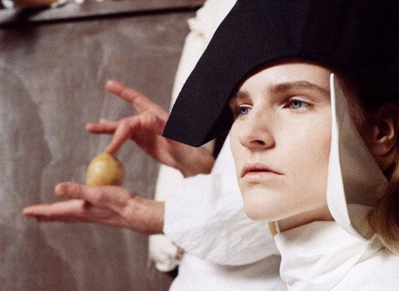 Close-up of a person wearing a structured black cap and white high-collar top, with another person holding a potato in the background