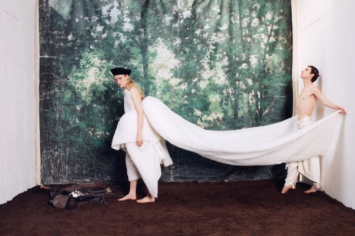 A woman in a white dress with a long train and a man holding it up against a painted forest backdrop.