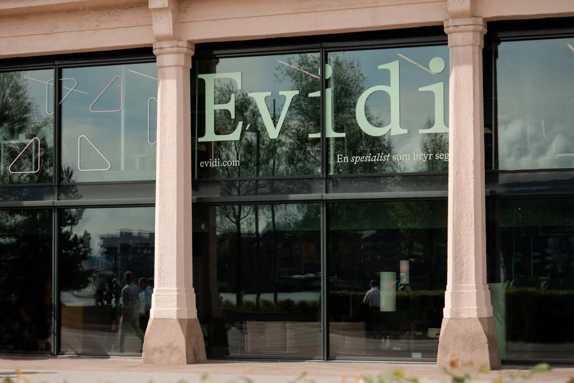 Exterior of a modern building with large glass windows displaying 'Evidi' logo and geometric shapes, reflecting trees and passersby.