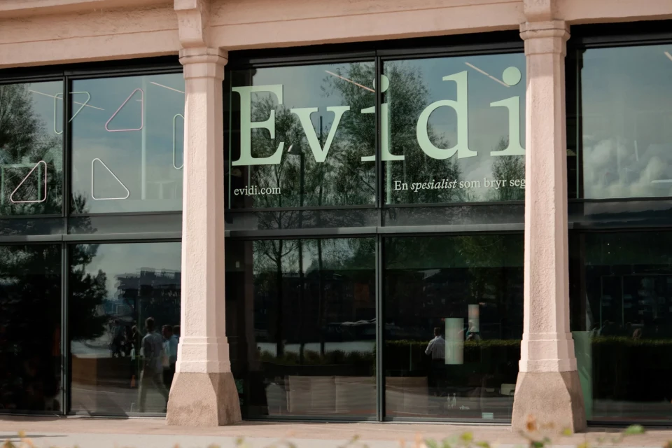 Exterior of a modern building with large glass windows displaying 'Evidi' logo and geometric shapes, reflecting trees and passersby.