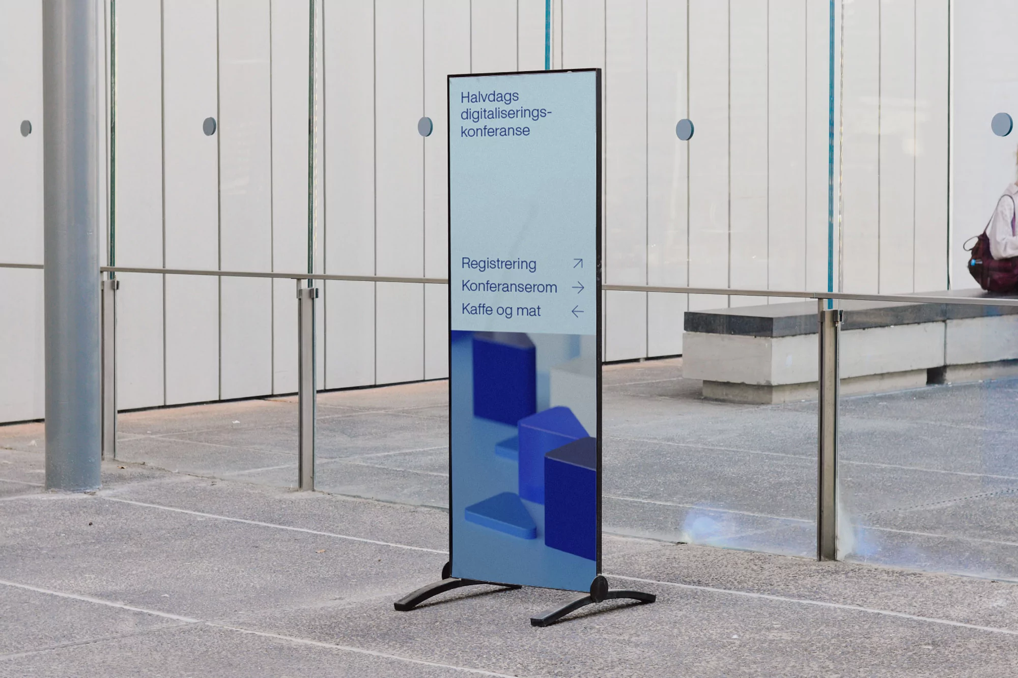 A vertical advertising banner with text and blue geometric designs on a grey floor inside an office building with white walls and metal railings.
