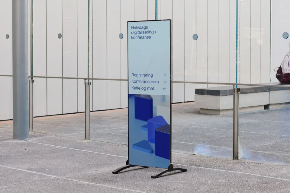 A vertical advertising banner with text and blue geometric designs on a grey floor inside an office building with white walls and metal railings.