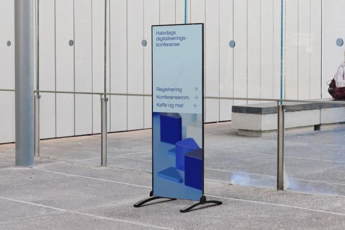 A vertical advertising banner with text and blue geometric designs on a grey floor inside an office building with white walls and metal railings.