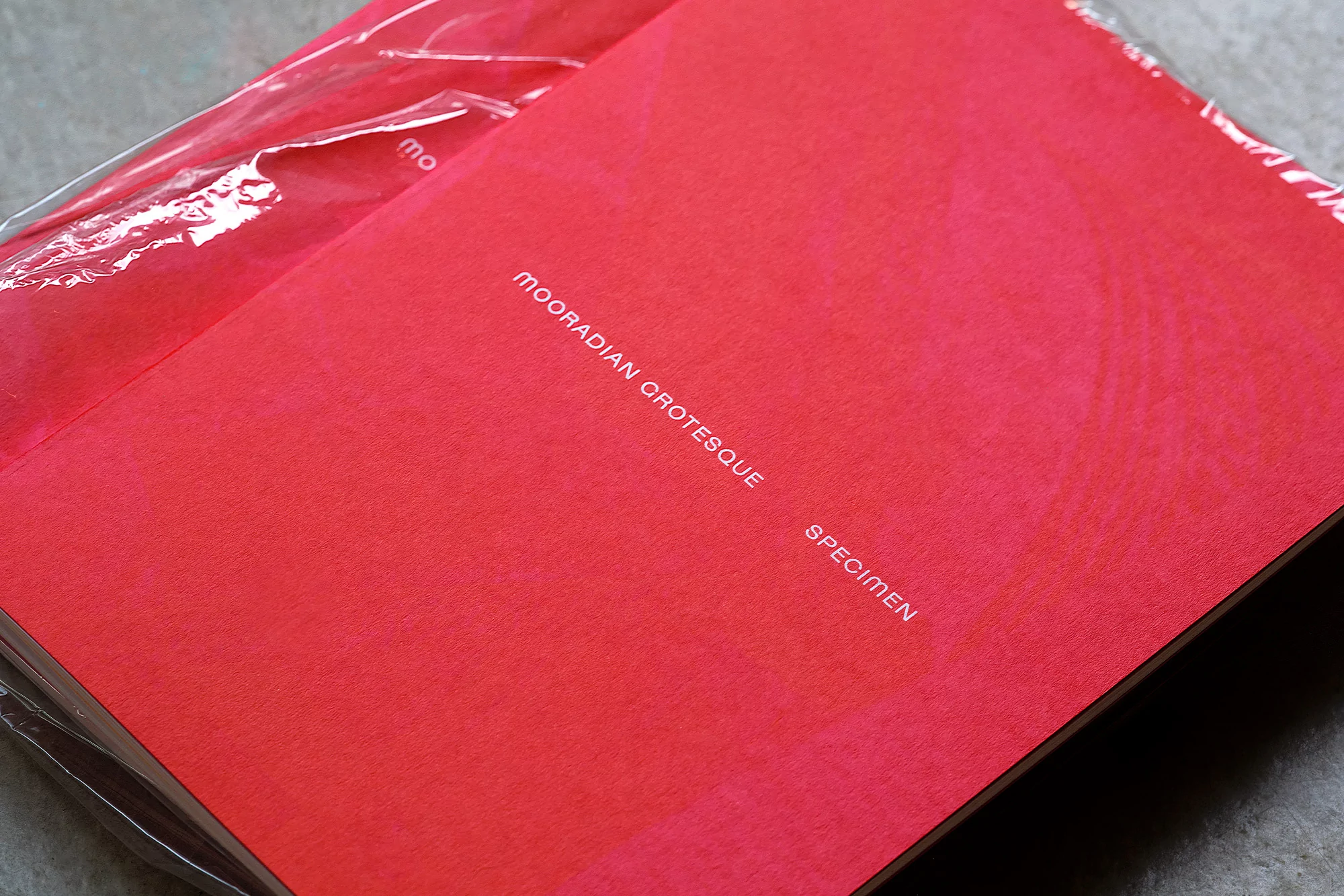Close-up of a red brochure or booklet with the text 'Mooradian Grotesque Specimen' and a clear plastic wrap, set on a gray surface.