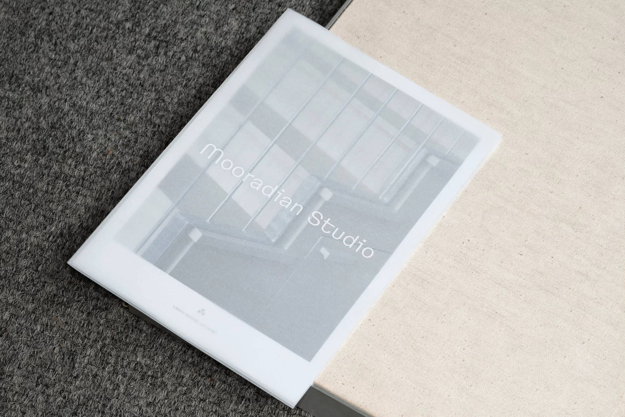 Close-up of a frosted translucent presentation book displaying architectural designs from Mooradian Studio, set against a gray carpet and beige linen.