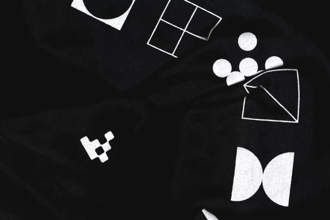 Black fabric background with scattered white geometric shapes including circles, squares, triangles, and other abstract designs.