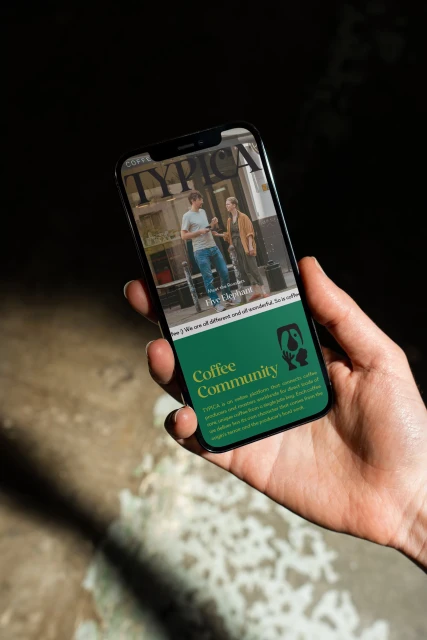 Hand holding a smartphone displaying a magazine cover with two people standing in front of a coffee shop entrance on the screen.