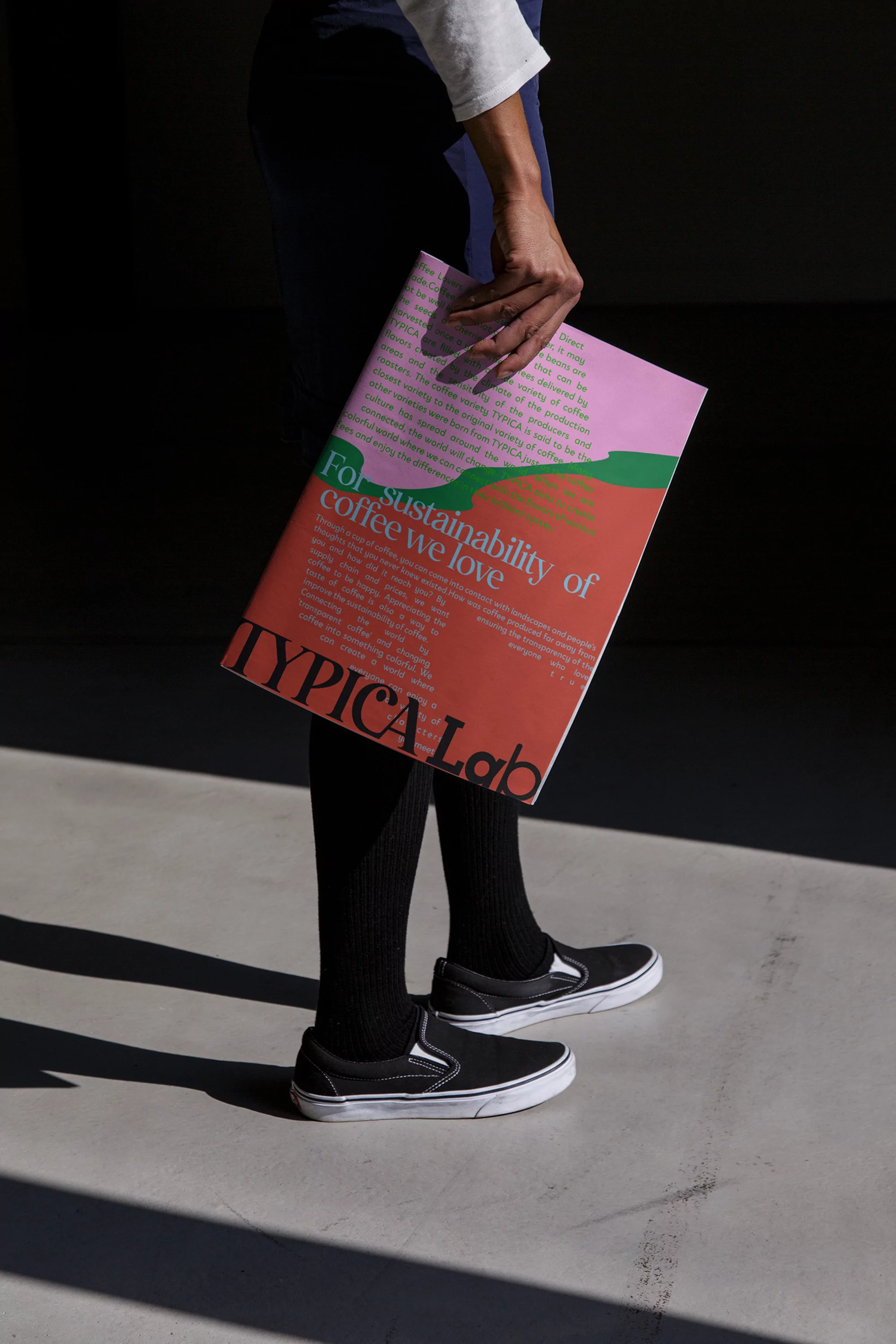 Person holding a magazine titled 'Typical Lab' with the text 'For sustainability of coffee we love', posing in a dimly lit indoor space.