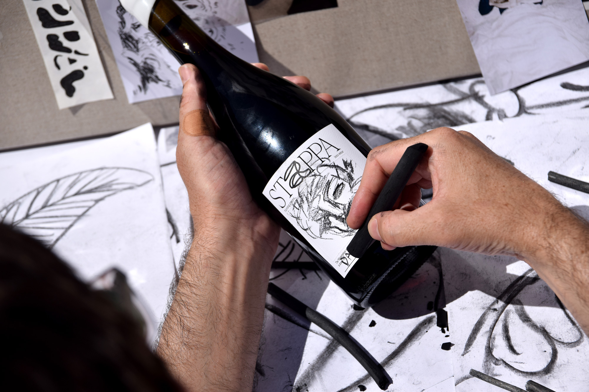 Person drawing on wine bottle label with charcoal, surrounded by paper drawings. Artistic expression merges wine with art.