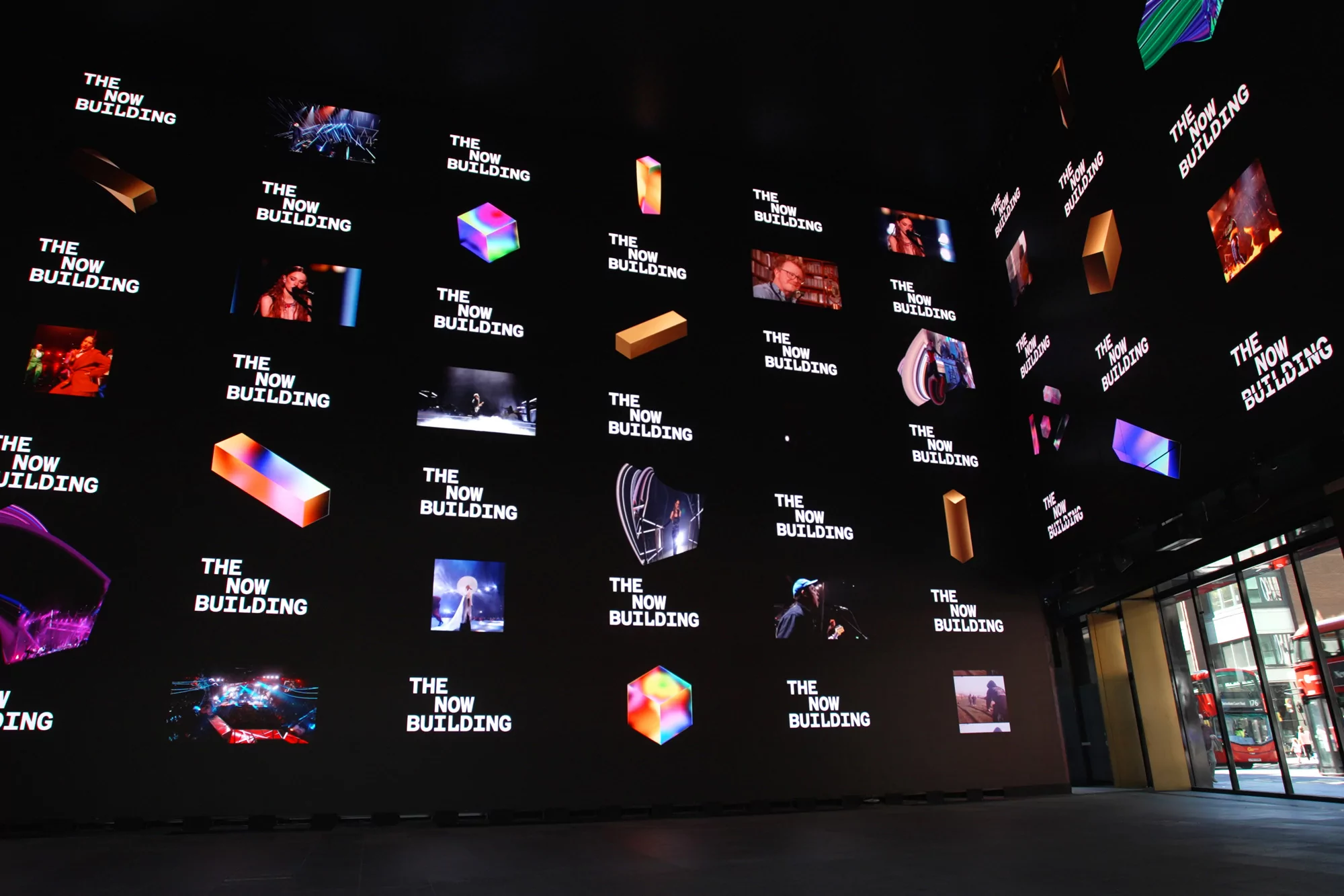 Indoor shot of an immersive display featuring the repeating brand text 'The Now Building' and abstract 3D designs, viewed from a low angle.