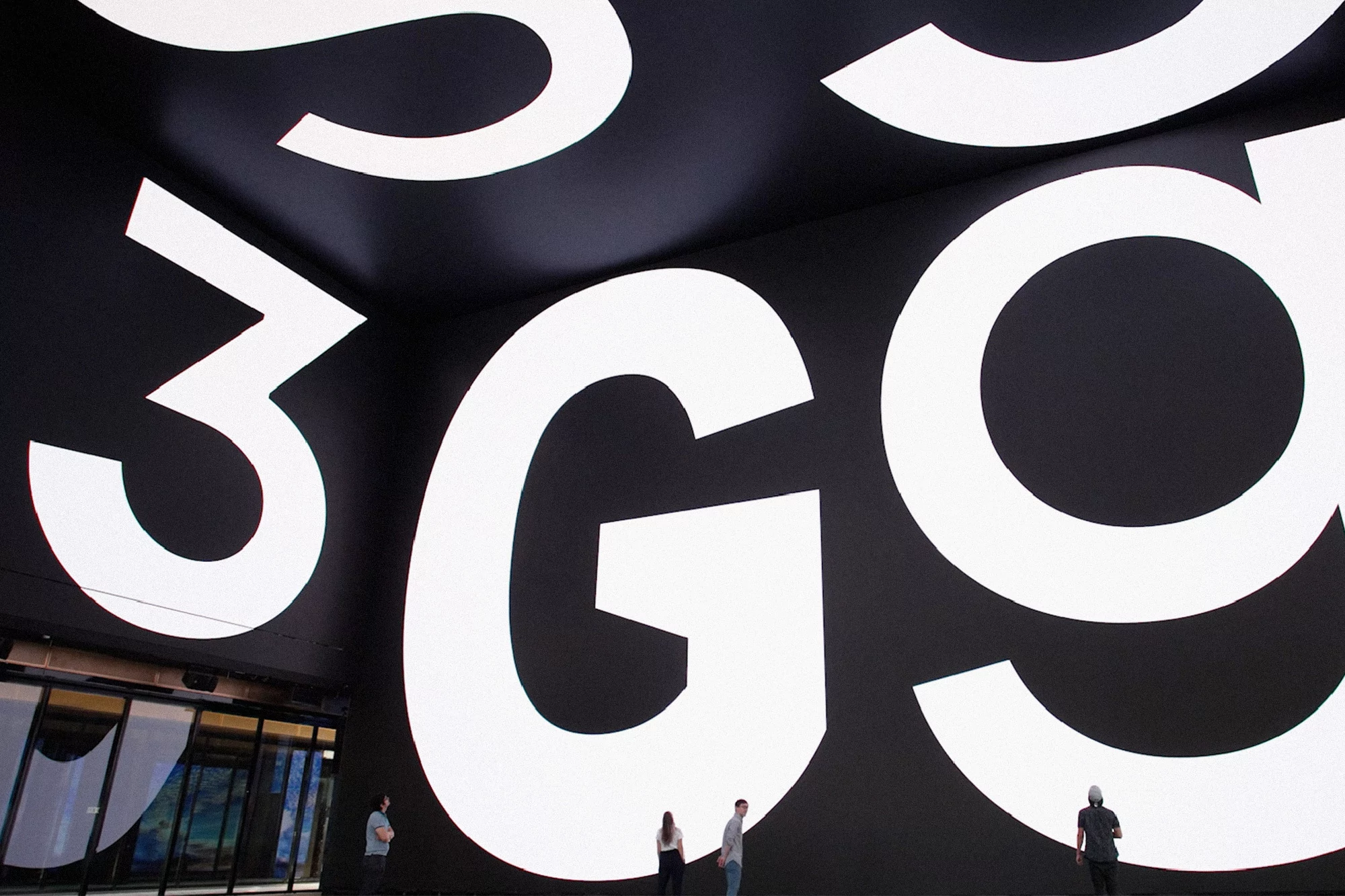 Large white 'G' and '3' typography on a black wall, four people standing nearby in an indoor setting, showcasing scale and design.