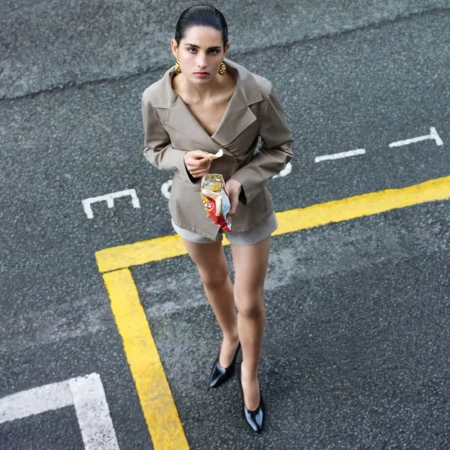 Woman in blazer and shorts stands on asphalt with painted lines, holds a snack, looking up at an overhead camera angle.