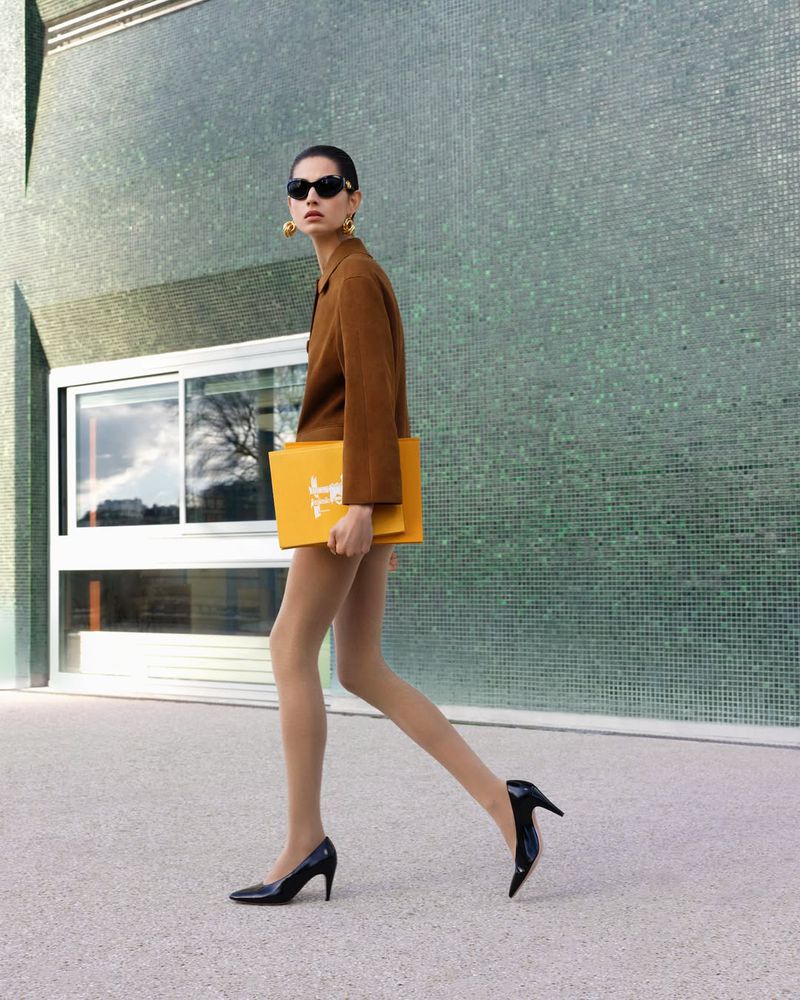A woman in a brown jacket and black heels walks past a textured green wall, carrying a yellow document case in a fashion setting.