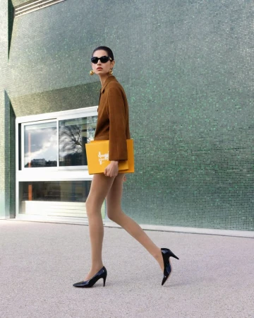 Model Africa Peñalver walking in a brown jacket and black heels carrying a yellow folder against a green tiled wall.