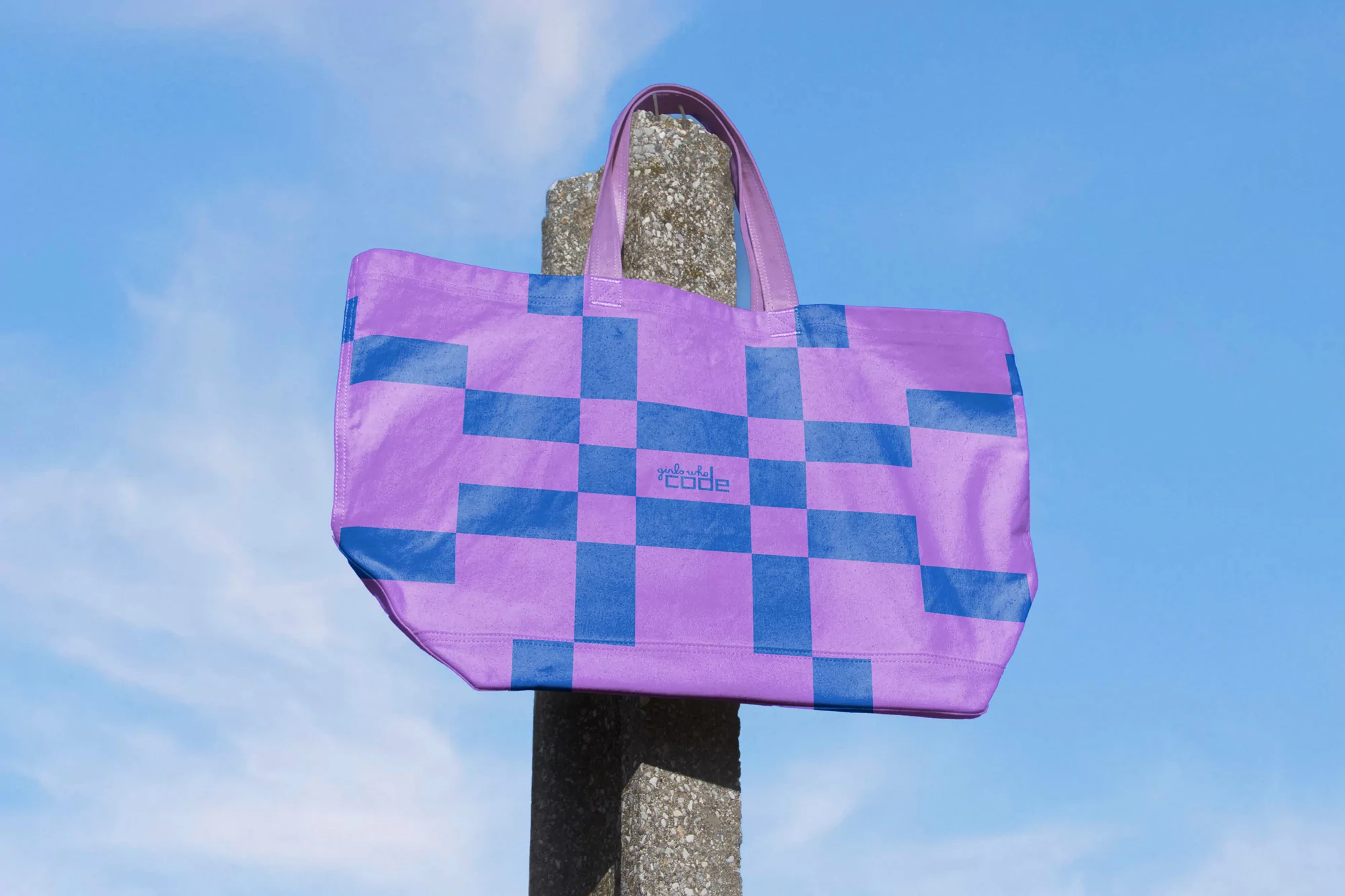 A purple tote bag with a blue pixelated square pattern hangs on a concrete post against a blue sky with sparse clouds.