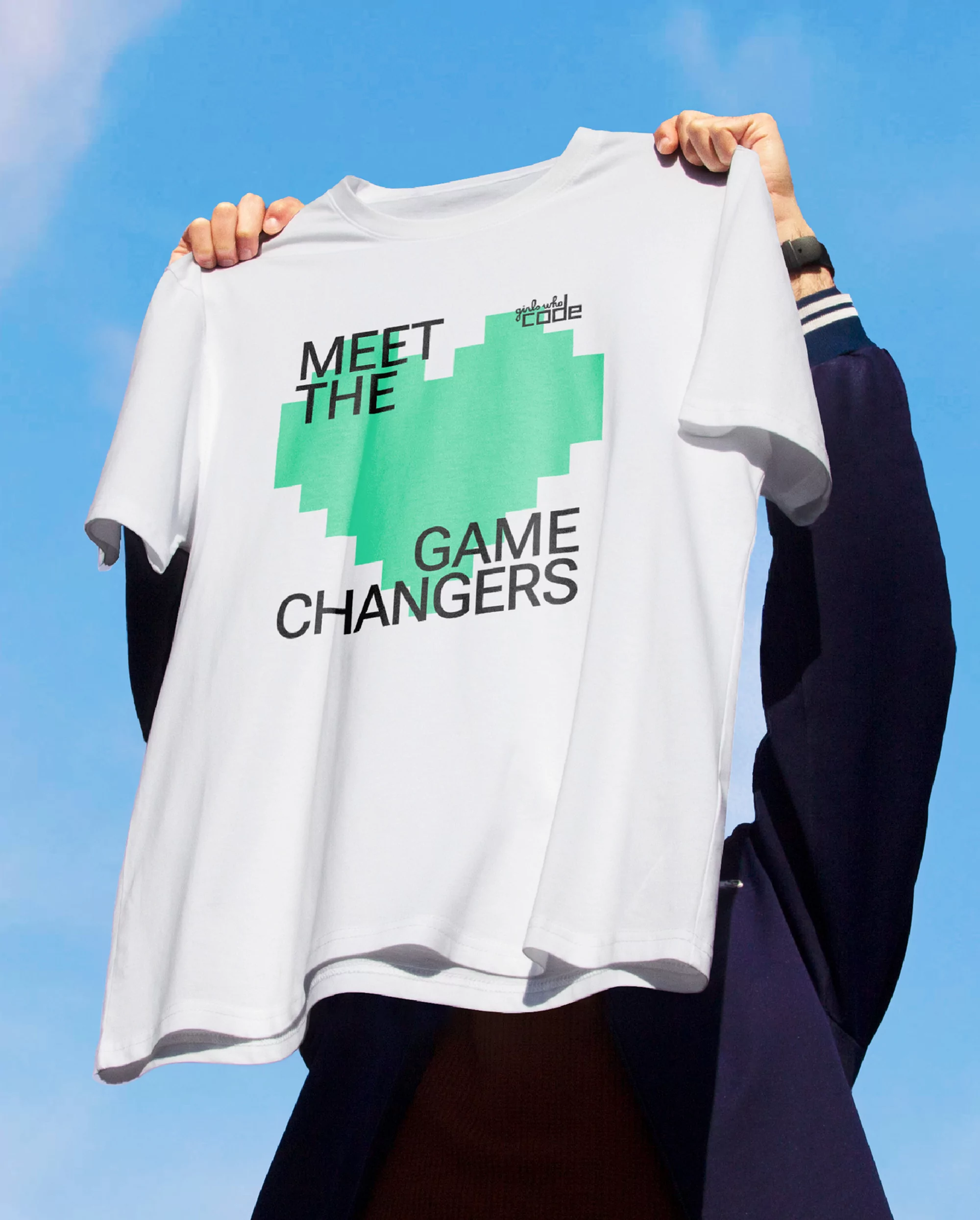 A person holds up a white t-shirt against a blue sky. The shirt features pixelated art and the text 'Meet the Game Changers'.