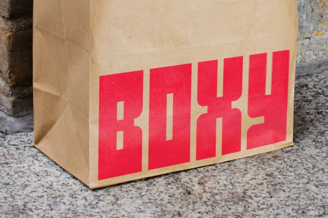 Close-up of a brown paper shopping bag with large red lettering that reads "BOXY" on a textured, grey concrete surface.