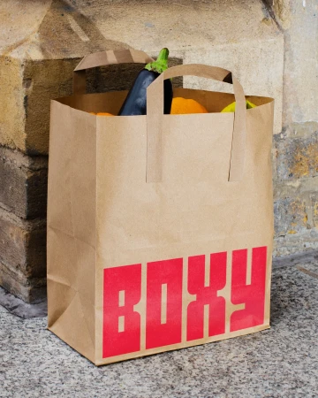Brown paper grocery bag with the word 'BOXY' in red letters, holding an eggplant and oranges, on a stone surface against a weathered wall.