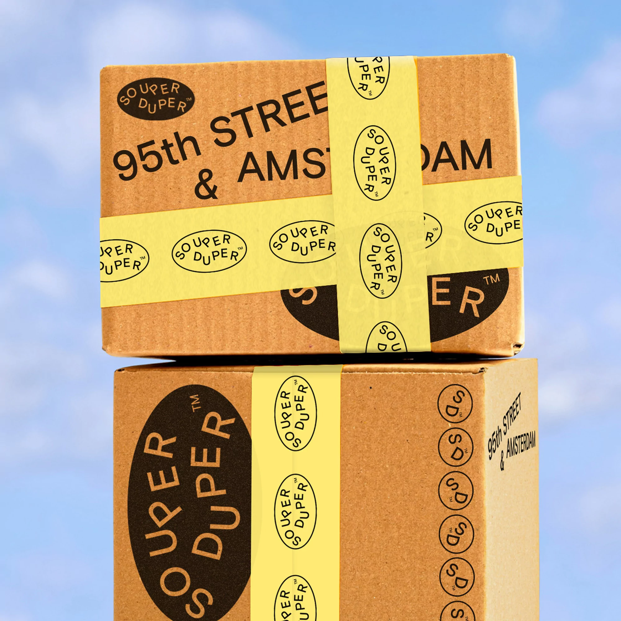 Two stacked cardboard boxes with yellow tape, featuring the 'Souper Duper' logo and '95th Street & Amsterdam' text against a blue sky backdrop.