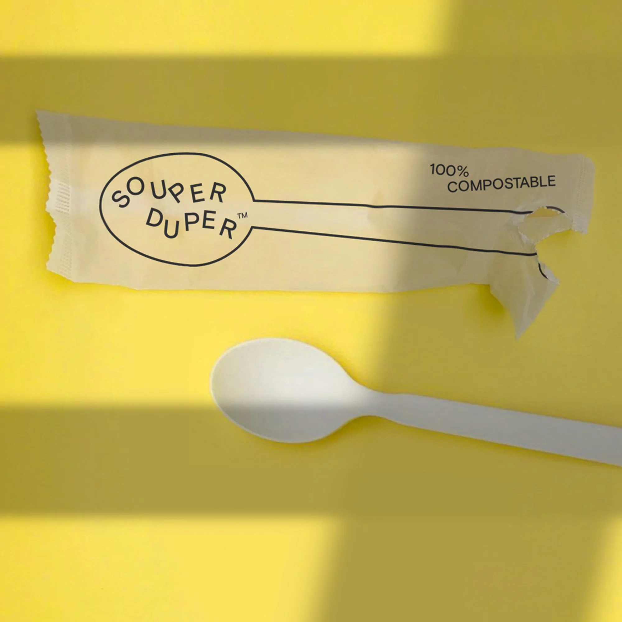 A compostable white plastic spoon and its wrapper, labeled Souper Duper, are on a yellow surface with shadows from sunlight.