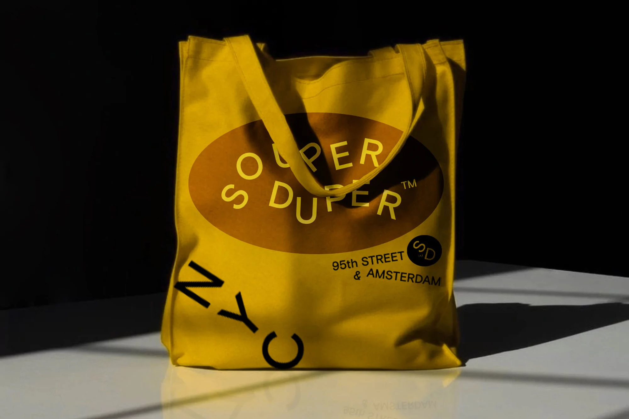 Yellow tote bag with brown oval logo. The tote bag says "Super Duper," "NYC," and "95th Street & Amsterdam." The bag sits on a white surface.