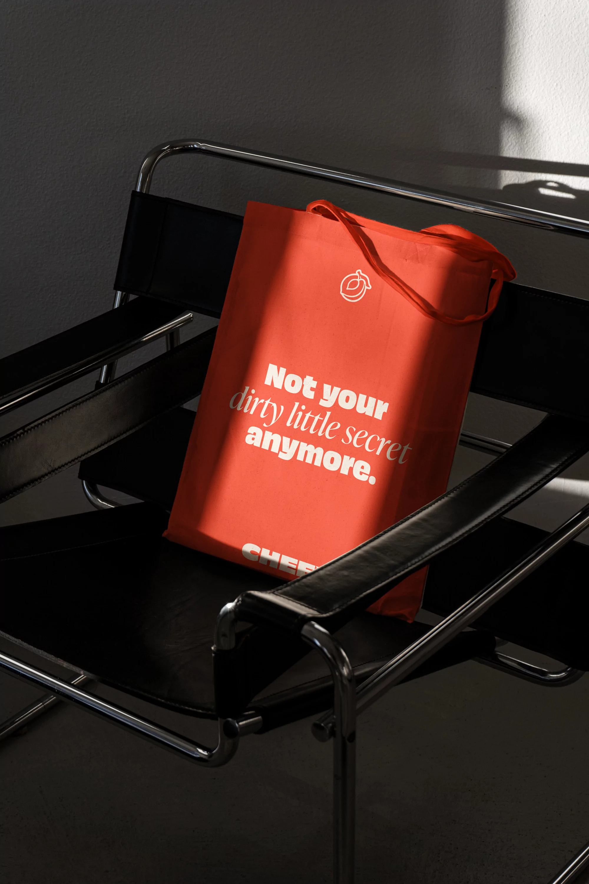 Orange canvas tote bag with a white slogan resting on a black Wassily chair indoors, bathed in sunlight creating strong shadows.