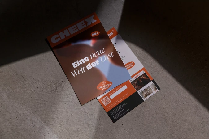 Two marketing flyers are displayed on a gray stone surface under hard, directional lighting creating shadows and promoting CHEEX.