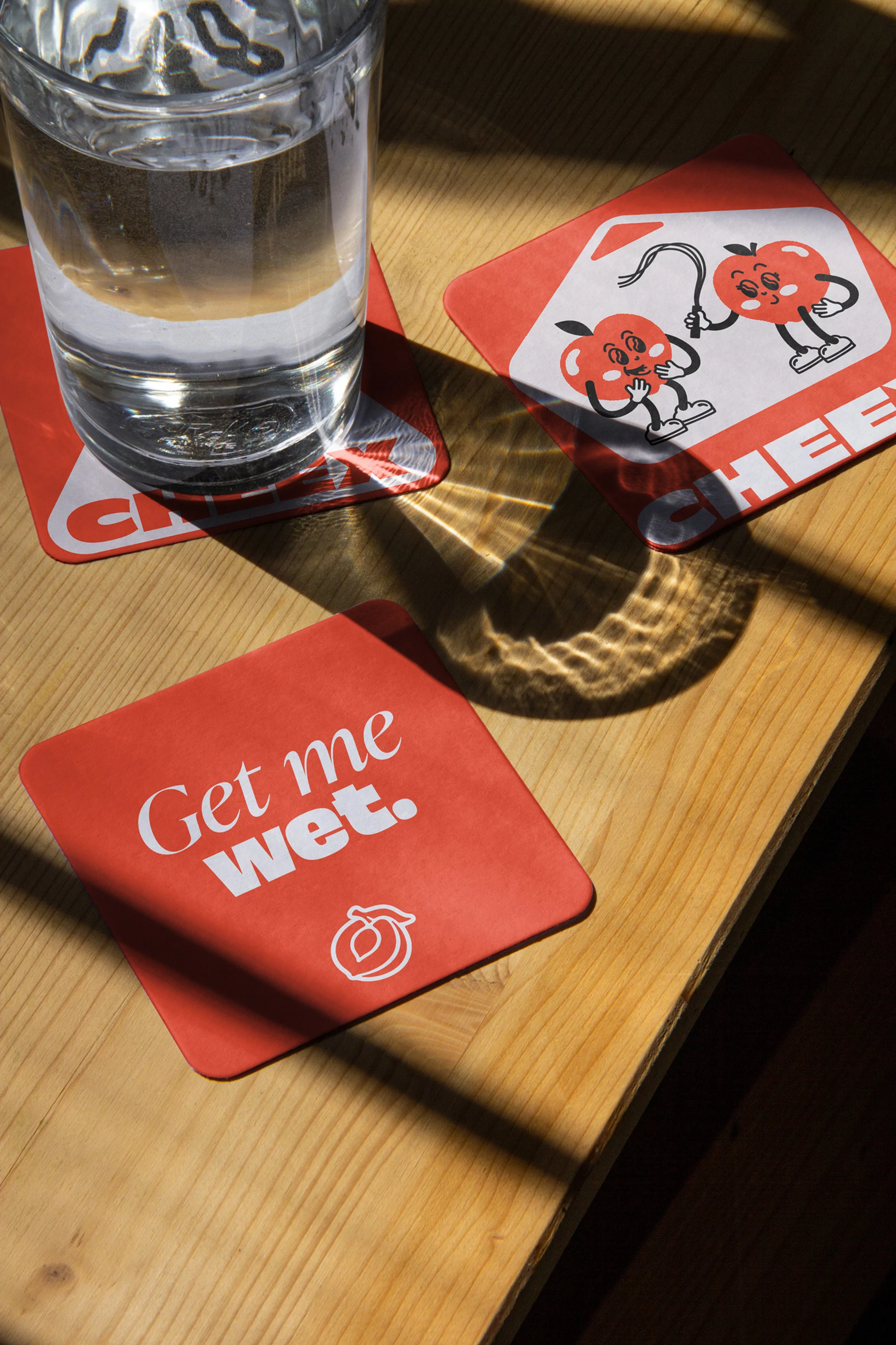 Glass of water and three square coasters on a wooden table illuminated by natural sunlight creating bold shadows across the surface.