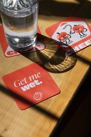 Glass of water and three square coasters on a wooden table illuminated by natural sunlight creating bold shadows across the surface.