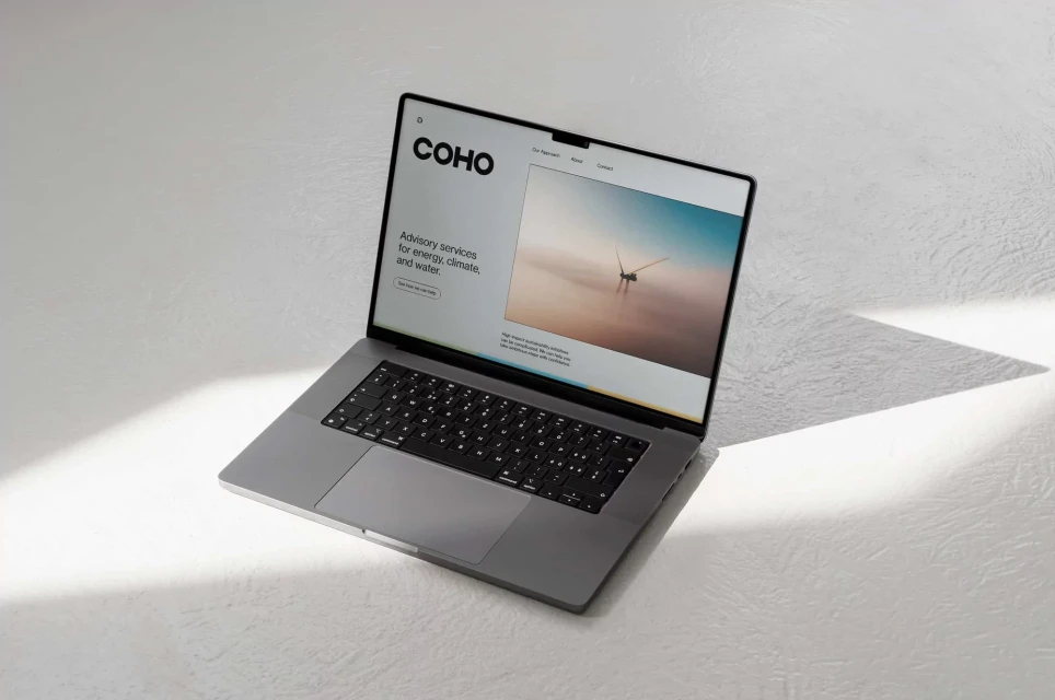 Laptop displaying COHO website on a white surface, showing advisory services for energy, climate, and water with a wind turbine image.