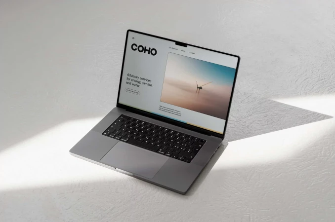 Laptop displaying COHO website on a white surface, showing advisory services for energy, climate, and water with a wind turbine image.