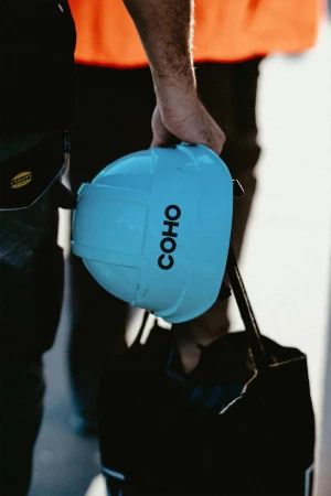 Construction worker holds a light blue hard hat with 'COHO' printed on the side, wearing black pants and an orange vest, mid-shot.