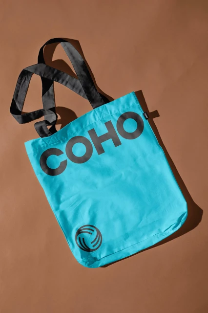 Turquoise tote bag featuring black handles and the word "COHO" in black letters on a tan background. Merchandise concept.
