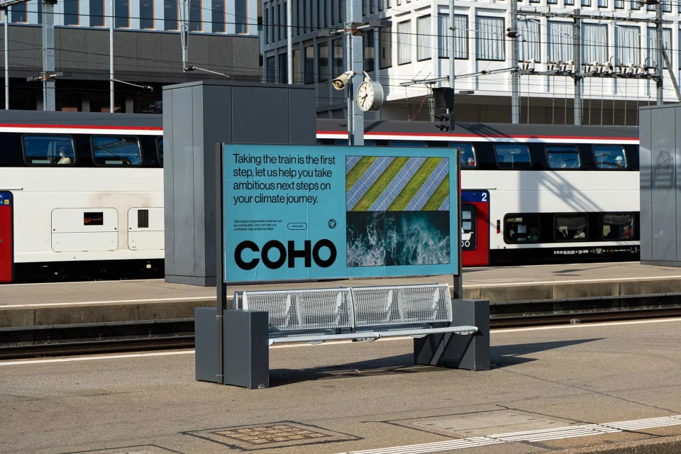 Billboard advertisement for COHO next to a train station bench with a passenger train passing by in the background.