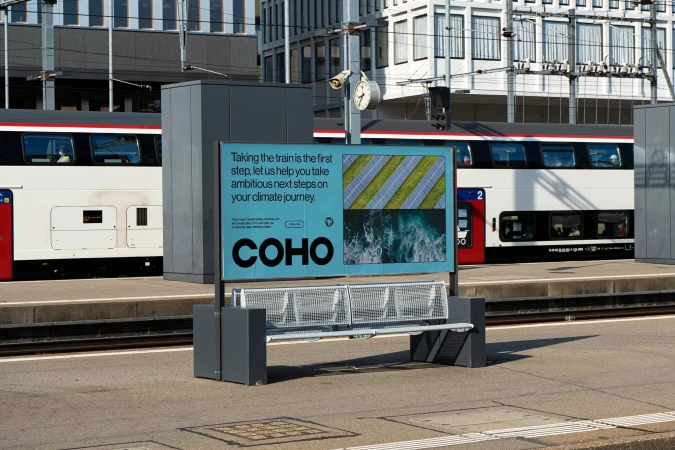 Billboard advertisement for COHO next to a train station bench with a passenger train passing by in the background.