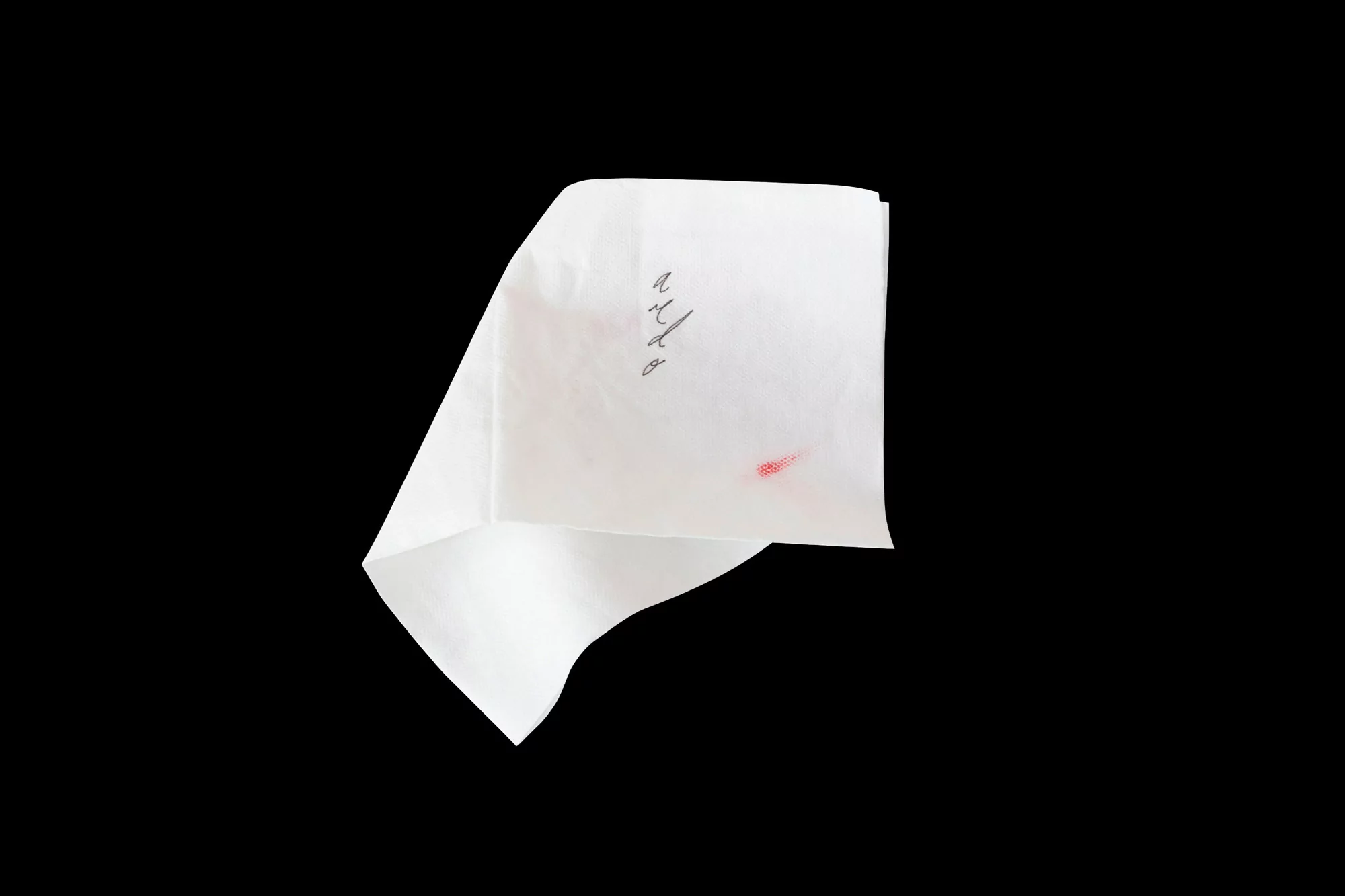 Close-up of a folded white paper with handwritten script and a small red stain against a stark black background, emphasizing texture and contrast.