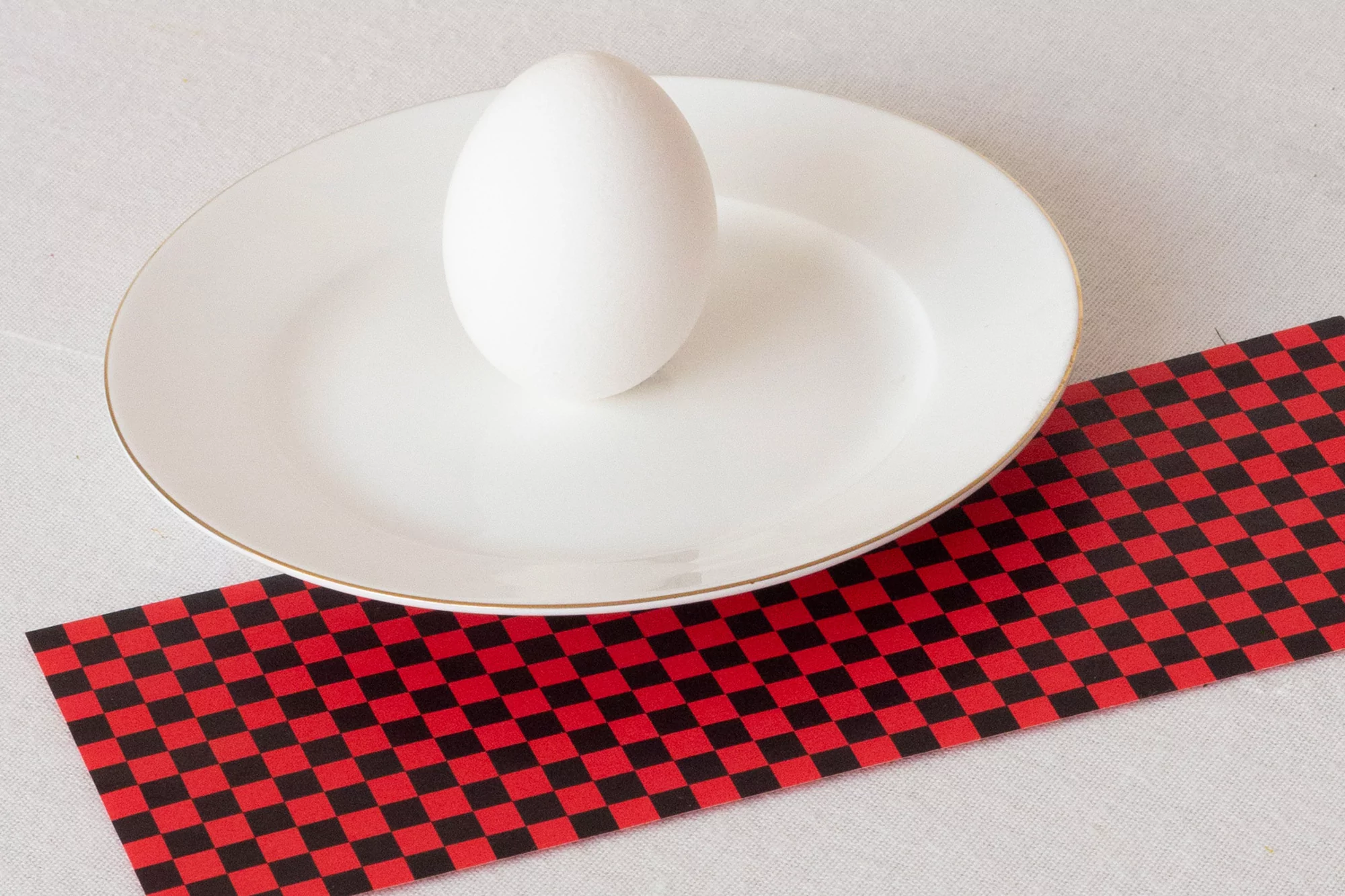 Elevated shot of a white egg resting on a white plate with a gold rim, placed on a white tablecloth with red and black checkered paper.