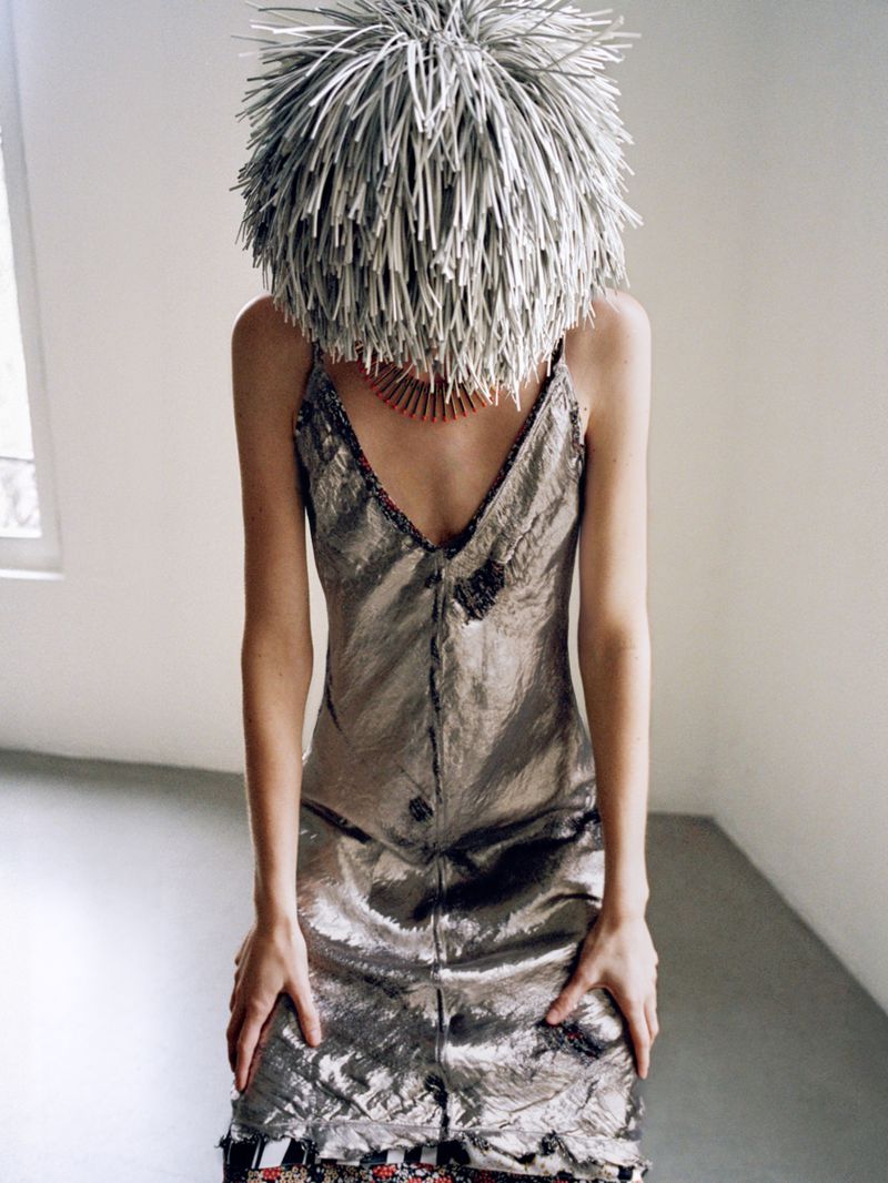 Woman in shimmering dress with decorative spiky headpiece stands in empty room with window. Soft natural lighting creates a muted ambiance.