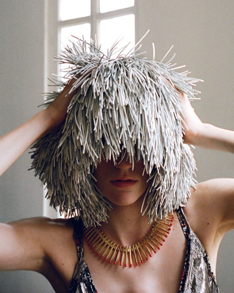Adult female wearing a spiky gray wig and a gold and red necklace standing indoors in front of a white window, fashion portrait. 