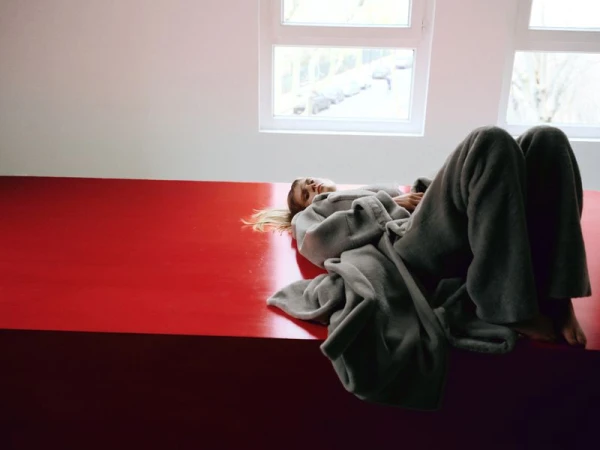 Blonde woman in grey robe lies on red platform in front of large windows, light indoor scene, relaxing at home.