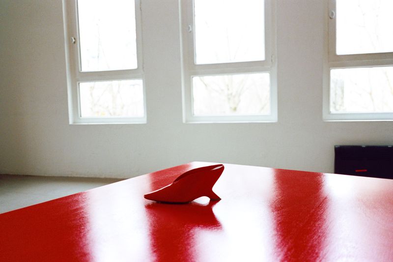 Red shoe rests on a red glossy table in a white room. The setting includes three windows and hints of outdoor trees and greenery.