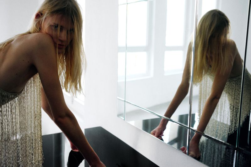 Blonde woman in a fringed top leans on a black table next to a large mirror in a bright room with white walls and natural light.