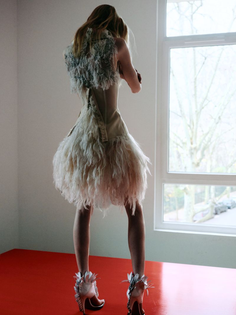 Blonde woman in a feathered mini-dress and red platform heels poses in front of a window, showcasing avant-garde fashion in a studio setting.