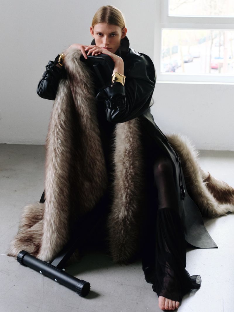 Elegant woman in black coat poses with a fur throw, sitting against a white wall near a window, showcasing fashion and style.