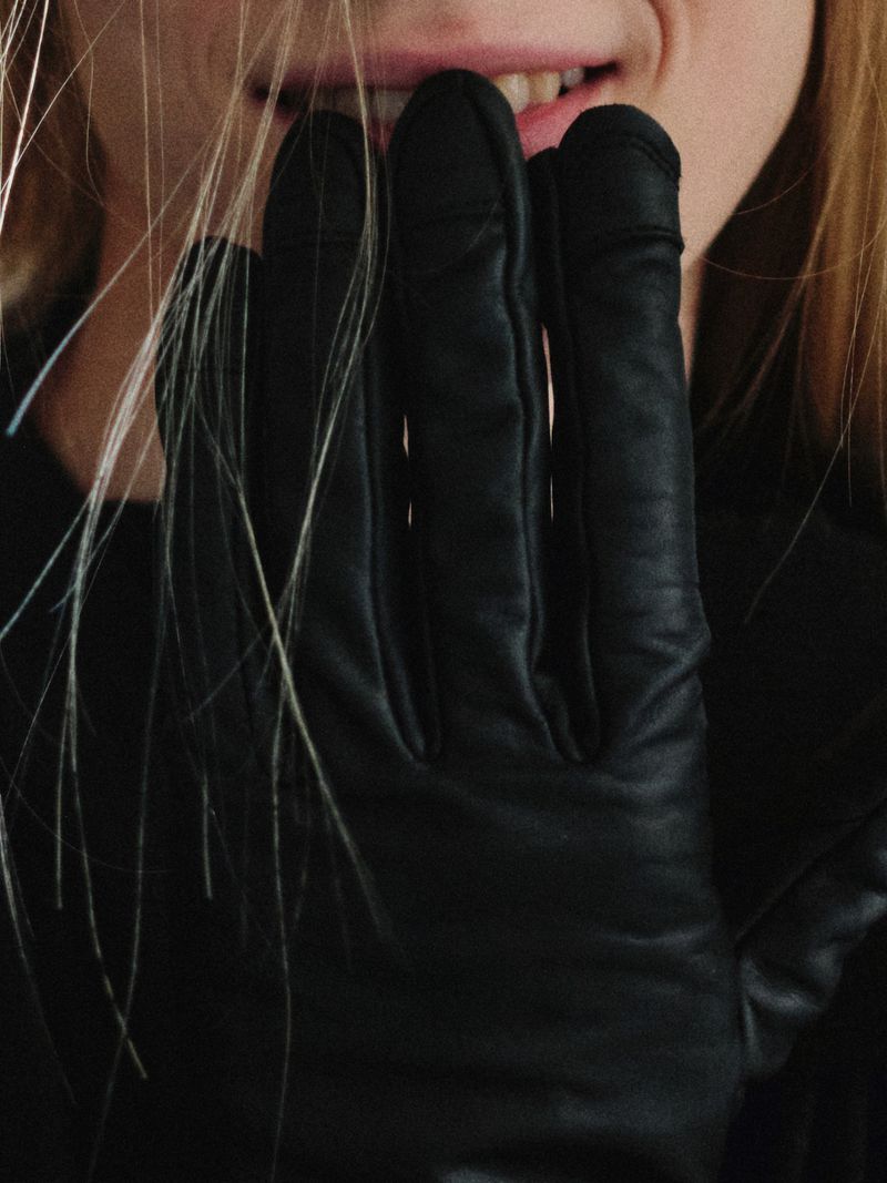 Close-up shot of a woman with blond hair wearing black leather gloves, fingertips touching her face near her lips with visible teeth.