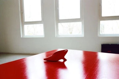 A small red sculptural object resting on a polished red table in front of bright windows.